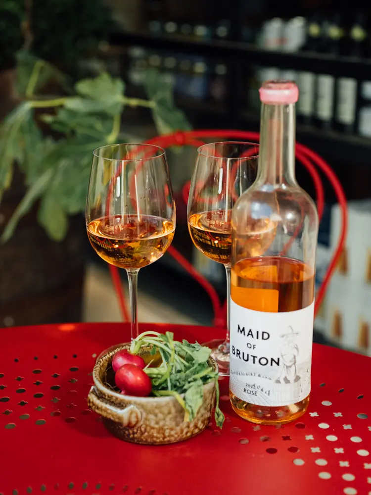 A table with a red tablecloth with two glasses of rose, a bottle of rose and a basket with radishes