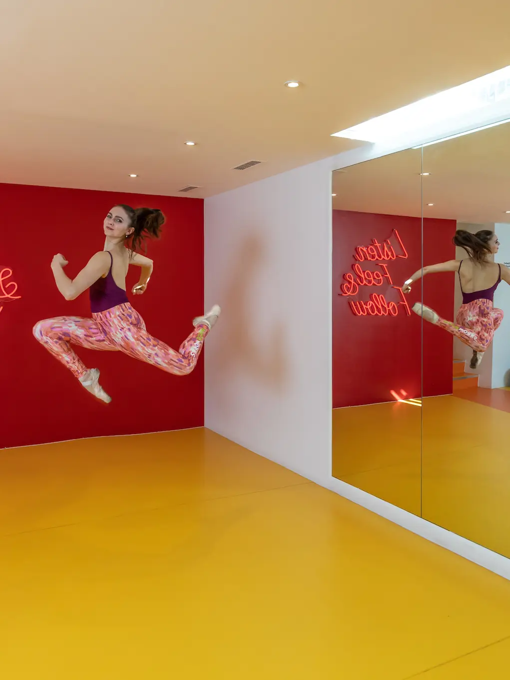 Dance studio with a yellow floor, red wall with artwork showing woman jumping in the air, and a mirror on the right wall