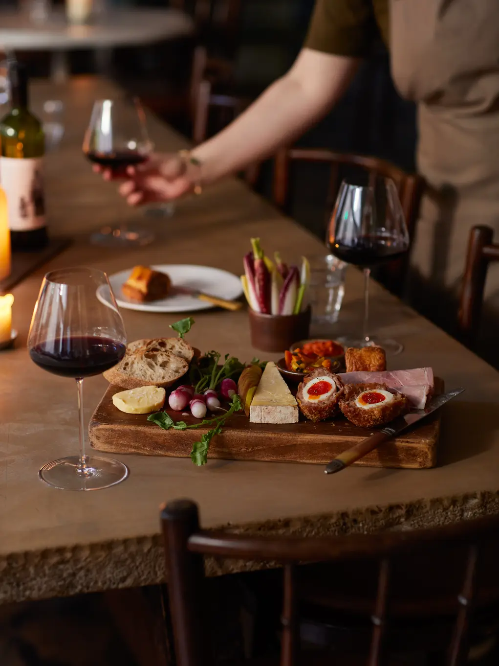 A wooden table with glasses of red wine and a wooden chopping board with Scotch eggs and other food